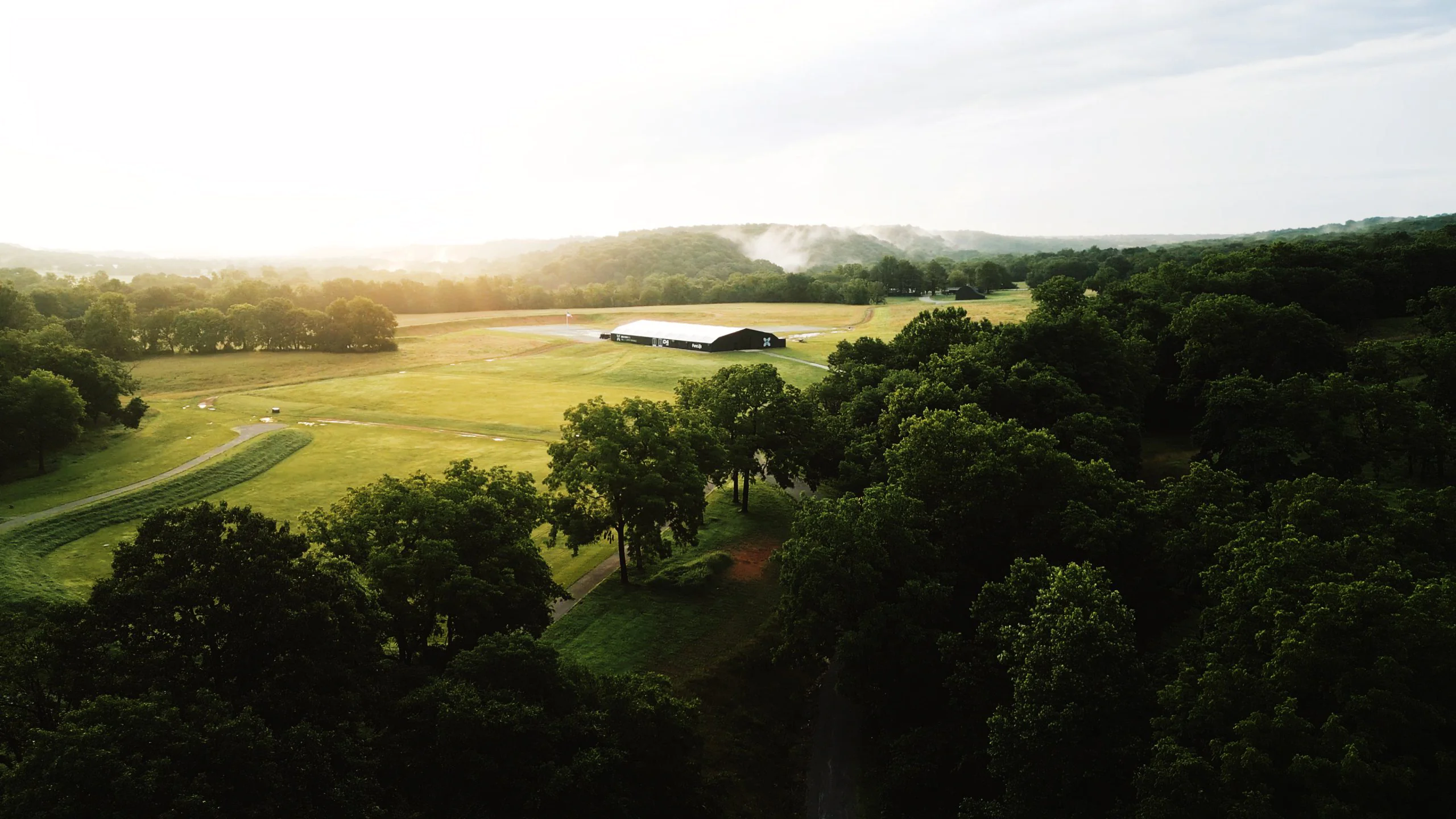 Aerial view of a green, tree-lined landscape with a large barn in the center, open fields, winding paths, and mist rising in the distance under a bright sky.