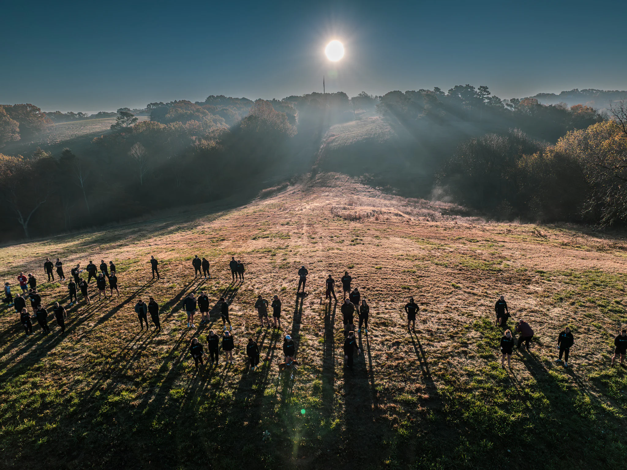 A group of people stand scattered across a sunlit field at sunrise, casting long shadows on the grass with trees, rolling hills, and a bright sun in the background.