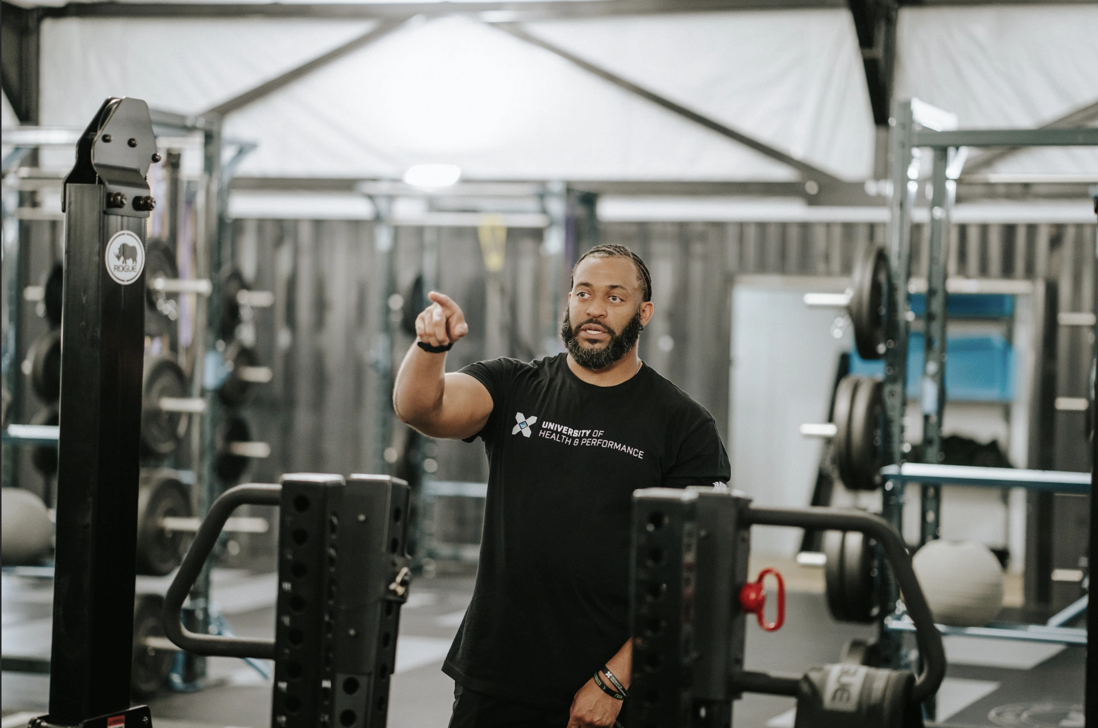 81351d373ed750e0f4a694bb56aef38cda89b83f A man wearing a black t-shirt stands in a gym surrounded by weightlifting equipment, gesturing with his right hand as if instructing or guiding someone.