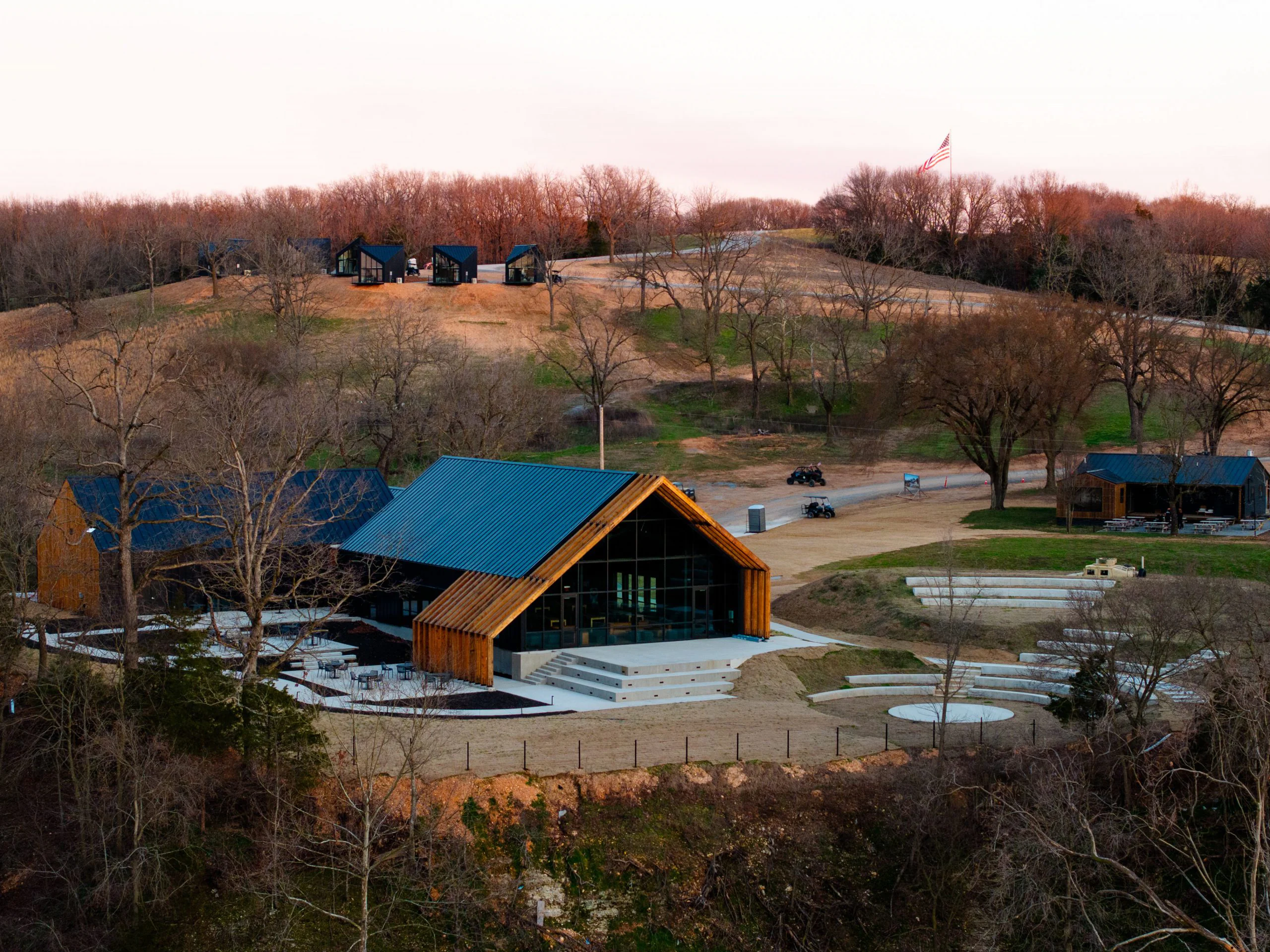 A modern building with a black roof and wood accents is surrounded by leafless trees and rolling hills. Small black cabins are scattered on a hill in the background, with an American flag visible at the top.