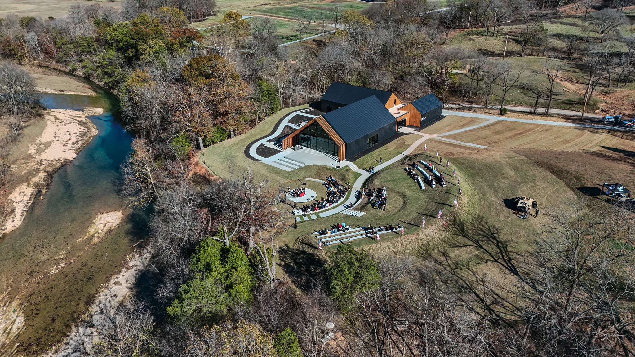 Aerial view of an outdoor wedding ceremony near a modern building by a river, surrounded by trees and open grassy fields. Guests are seated in rows facing a circular area where the ceremony is taking place.