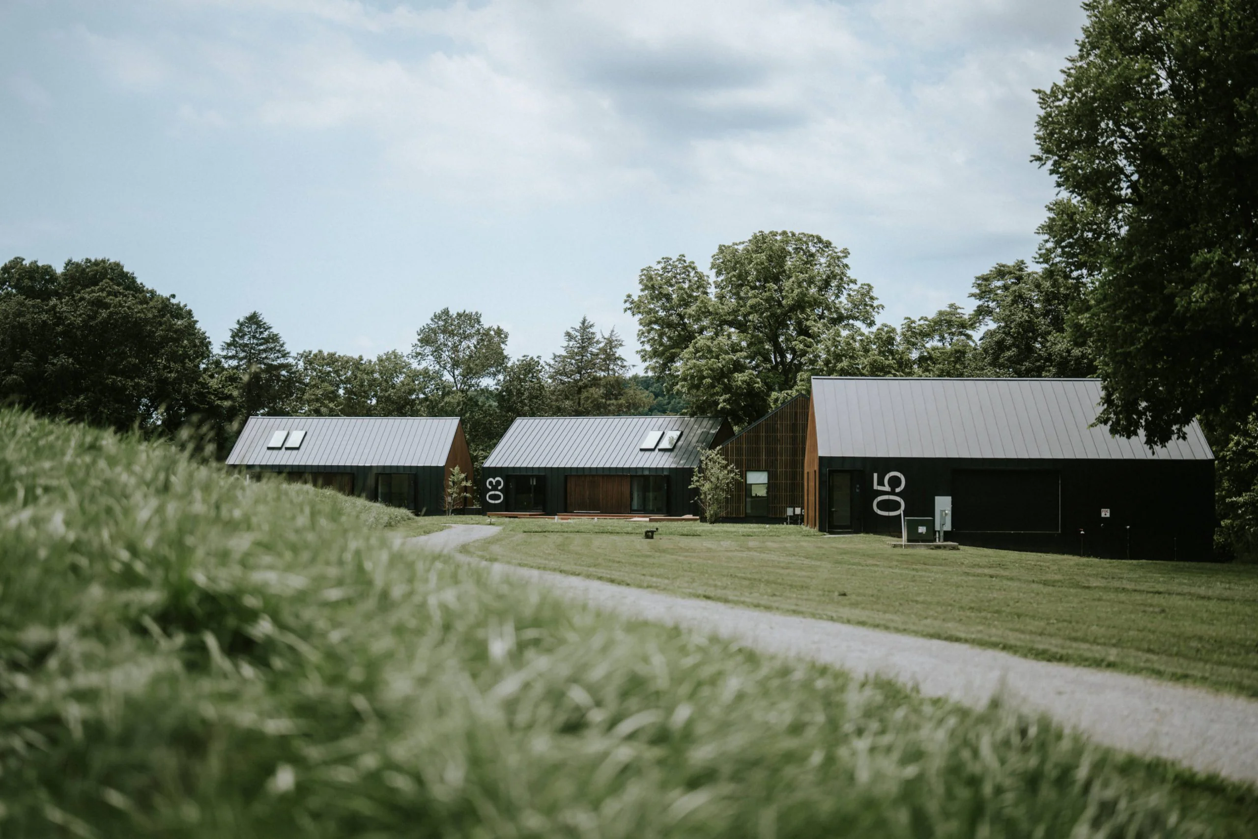 Three modern, black cabins with metal roofs sit on a grassy lawn near a winding path, surrounded by green trees. The cabins are labeled with large white numbers: 03, 04, and 05. The sky is partly cloudy.