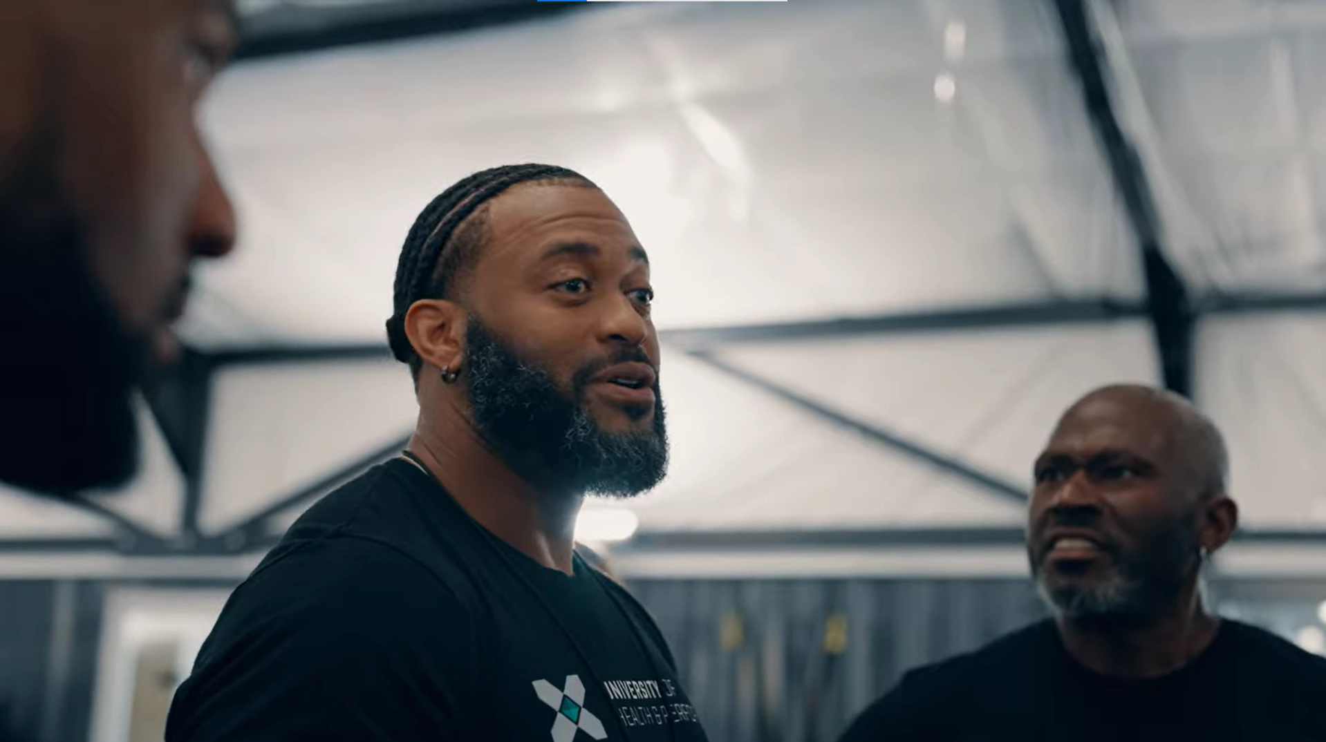 Three men with beards stand indoors, engaged in conversation. The central man speaks, wearing a black shirt with a logo, while the man on the right looks on, appearing focused. The background shows a high-ceilinged, industrial space.