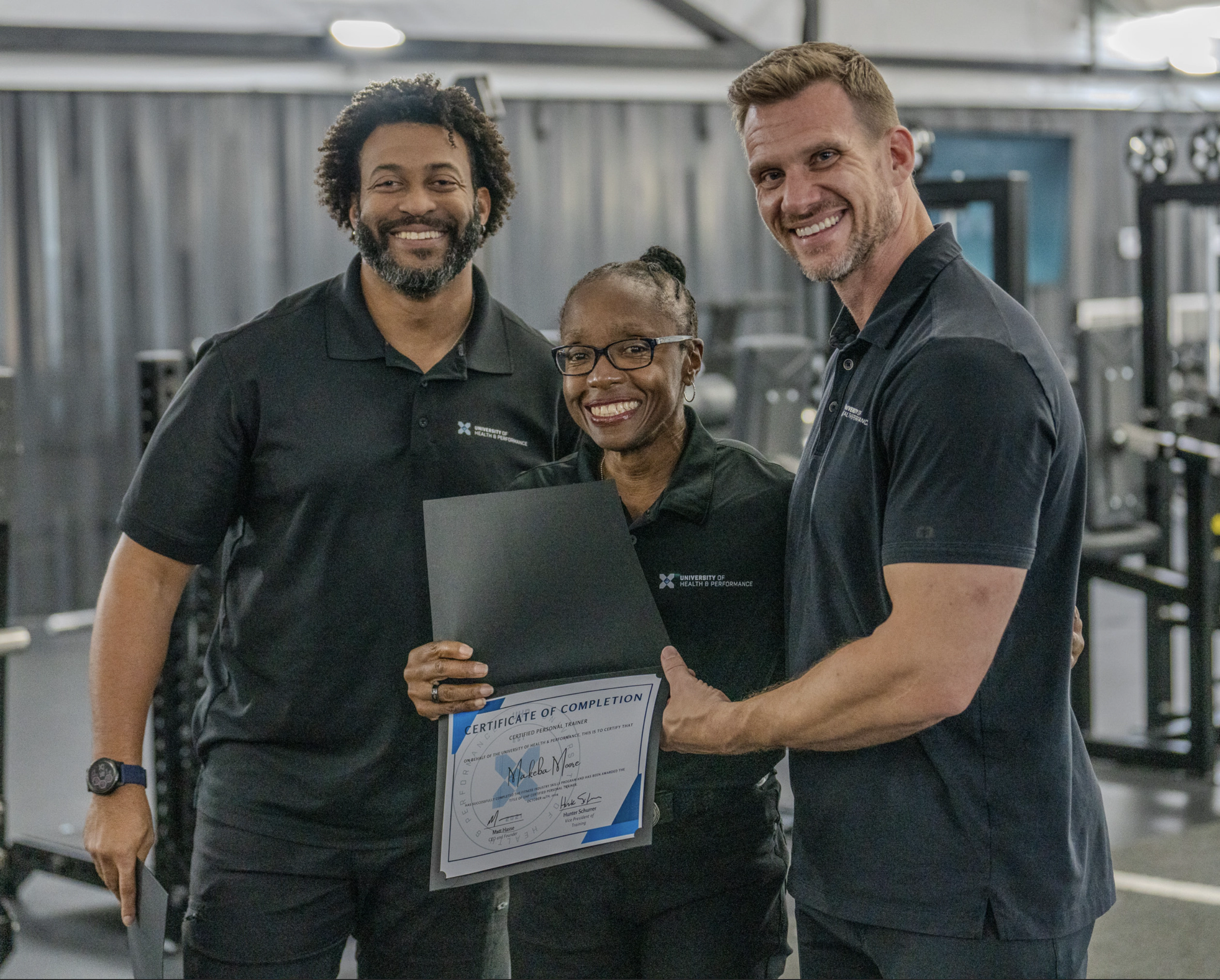Three people standing together in a gym, smiling. The person in the center holds a certificate of completion, while the other two stand on either side, celebrating the achievement. All are wearing black polo shirts.