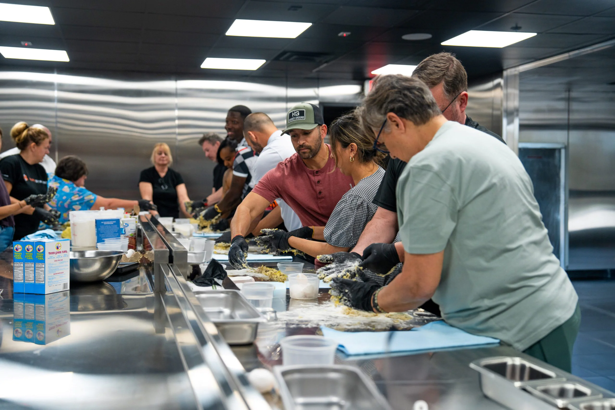 ff89dbf2615ba022714ffd2b8bac8ea62f7e194e A group of people work together in a modern kitchen, preparing food at a long counter. Some are mixing dough by hand, while others measure ingredients; various bowls and supplies are scattered across the workspace.